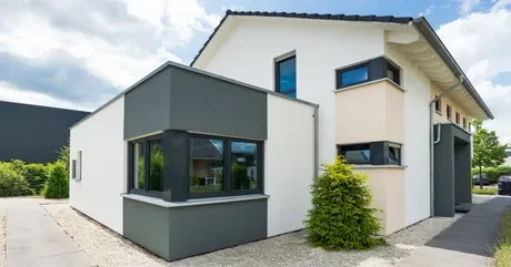 Maison moderne à façade blanche et grise, avec des fenêtres angulaires et vegetation autour.